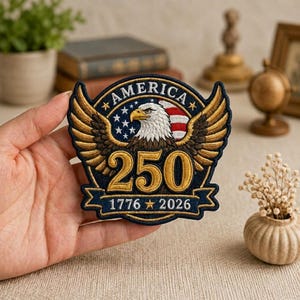 May include: A circular embroidered patch featuring a bald eagle with spread wings, a blue background with white stars, and red and white stripes. The text reads AMERICA, 250, and 1776-2026 with gold stars. The patch has a dark blue border.
