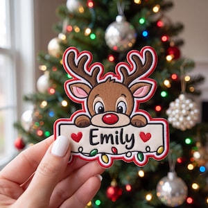 May include: A custom embroidered patch featuring a brown reindeer with a red nose, holding a rectangular sign that reads Emily in black text, flanked by two red hearts and decorated with colorful Christmas light bulbs along the bottom edge.