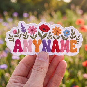 May include: A white embroidered patch featuring the word ANYNAME in colorful, rounded, puffy-style letters. Above each letter is a small, detailed floral stem with various blooms including pink, purple, red, orange, and blue flowers on green stems.