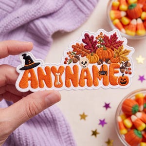 May include: An embroidered patch featuring the text ANYNAME in orange bubble letters. The design includes a black witch hat, a white bone, a small skull, a spider, and a jack-o'-lantern, all surrounded by colorful autumn leaves and acorns.
