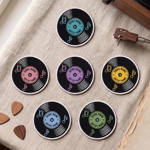 May include: Six circular coasters designed to look like vinyl records with black grooves and colorful center labels in teal, pink, purple, yellow, light blue, and green. Each label features the printed text SONG NAME and ARTIST NAME.