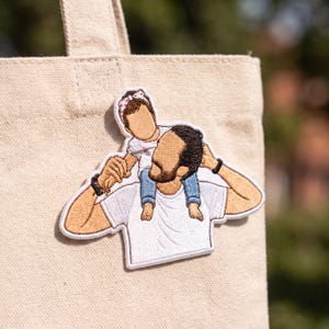 May include: An embroidered patch featuring a faceless figure carrying a small child on their shoulders. The figure wears a white t-shirt, while the child wears a white top and blue pants with a patterned headband. The patch is attached to a beige canvas tote bag.