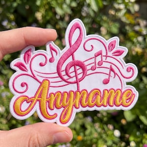 May include: A white embroidered patch featuring a large pink treble clef, musical notes, and swirling decorative lines. The word Anyname is stitched in a bold, golden-yellow script font across the bottom center of the white fabric base.