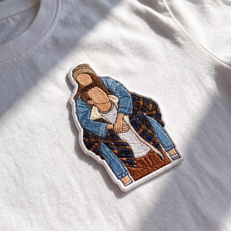May include: A white t-shirt featuring an embroidered patch of two figures. One figure wears a tan beanie, blue denim jacket, and brown pants. The other figure wears a dark blue and brown plaid flannel shirt over a white shirt and blue jeans.