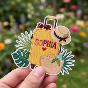 May include: An embroidered patch featuring a yellow suitcase with the name SOPHIA and a red heart, a straw hat, red sunglasses, a beige tote bag, and green tropical palm and monstera leaves. The design is outlined in white on a fabric base.
