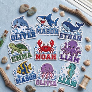 May include: A collection of nine die-cut stickers featuring cartoon sea creatures including a dolphin, shark, whale, turtle, crab, octopus, fish, jellyfish, and seahorse, each paired with a personalized name in bold, colorful block lettering.
