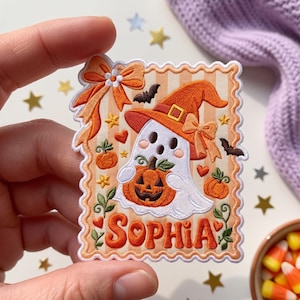May include: A rectangular embroidered patch with a scalloped edge featuring a white ghost wearing an orange witch hat and holding a jack-o'-lantern. The background has orange stripes, a bow, bats, and the name SOPHIA in orange lettering.