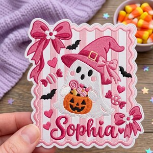 May include: A rectangular embroidered patch with a scalloped edge featuring a white ghost wearing a pink witch hat and holding an orange jack-o'-lantern bucket filled with candy. The background has pink stripes, pink bows, bats, hearts, and candies. The name Sophia is embroidered in pink script at the bottom.