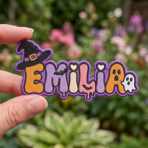 May include: An embroidered patch featuring the name EMILIA in a colorful, bubbly font with a purple border. The letter E is topped with a black witch hat, the M features a small black bat, the I is a heart, and the A includes a white ghost.