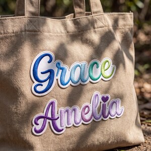 Peut inclure: Un sac fourre-tout en toile beige avec deux &eacute;cussons brod&eacute;s &agrave; bordure blanche. L'&eacute;cusson sup&eacute;rieur affiche le pr&eacute;nom Grace en &eacute;criture cursive d&eacute;grad&eacute;e du bleu au vert. L'&eacute;cusson inf&eacute;rieur affiche le pr&eacute;nom Amelia en d&eacute;grad&eacute; violet avec un c&oelig;ur.