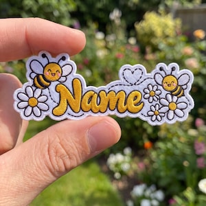 May include: An embroidered patch featuring the word Name in yellow script with a dark outline. The design includes two smiling bees with yellow and black stripes, white daisy flowers with yellow centers, and a small white heart outline.
