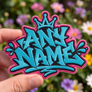 May include: An embroidered patch featuring the text ANY NAME in a bright cyan graffiti-style font with a black outline. The design is topped with a cyan crown and framed by a vibrant pink border, accented with small cyan droplet shapes on sides.