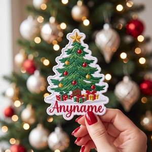 May include: An embroidered patch shaped like a Christmas tree with green branches, a brown trunk, a gold star topper, and red and gold ornaments. Beneath the tree are wrapped gifts and the red cursive text Anyname on a white background.