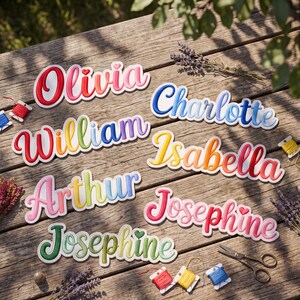 Peut inclure: Une collection d'&eacute;cussons brod&eacute;s portant les pr&eacute;noms Olivia, Charlotte, William, Isabella, Arthur et Josephine dans diverses couleurs et polices cursives, accompagn&eacute;s de bobines de fil, de ciseaux et de brins de lavande s&eacute;ch&eacute;e.
