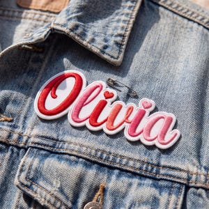 Peut inclure: Un &eacute;cusson brod&eacute; personnalis&eacute; affichant le pr&eacute;nom Olivia en &eacute;criture cursive avec des fils de couleur rouge, rose et blanc. L'&eacute;cusson comporte deux petits c&oelig;urs et est fix&eacute; sur une veste en jean bleu clair avec une &eacute;pingle &agrave; nourrice en m&eacute;tal argent&eacute;.
