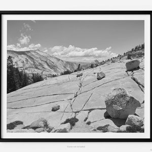 Puede incluir: Una fotografía en blanco y negro de un paisaje rocoso con una cordillera en el fondo. Las rocas están agrietadas y erosionadas, y hay varias rocas grandes dispersas por la superficie. El cielo está nublado y el sol brilla.