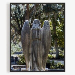 May include: A stone angel statue with wings, standing in a cemetery with trees and greenery in the background.
