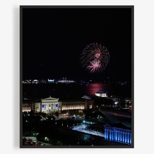 May include: A nighttime aerial view of a city skyline with fireworks exploding in the sky above a building. The fireworks are red, white, and blue.