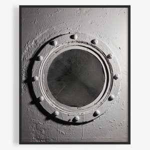 May include: A round metal porthole with a black circular window, surrounded by a ring of rivets. The porthole is set into a white painted wall.