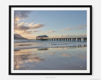 May include: A wooden pier extends out into the ocean at sunset. The sky is a soft blue with fluffy white clouds. The water is calm and reflects the sky and clouds. The beach is sandy and empty.