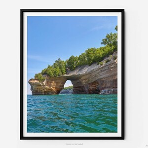 May include: A natural stone archway carved into a cliff face, with lush green trees growing on top. The archway is over a body of clear blue water.