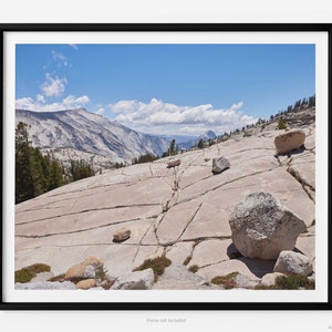 Puede incluir: Una vista panorámica de un paisaje montañoso rocoso con un cielo azul y nubes blancas. En primer plano, grandes rocas lisas y de color claro se encuentran dispersas sobre una superficie agrietada de color marrón claro.
