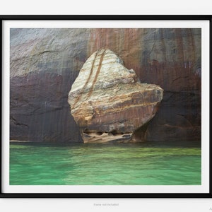 May include: A framed print of a rock formation emerging from turquoise water. The rock has layers of tan, brown, and white. The surrounding cliff is dark brown and gray. The image is framed in a black border.