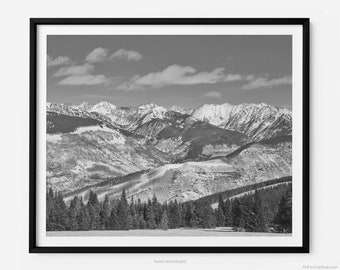 May include: A black and white photograph of a snow-capped mountain range with a forest in the foreground. The sky is partly cloudy.