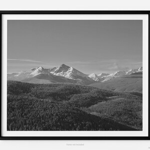 May include: A black and white photograph of a mountain range with snow-capped peaks. The mountains are in the distance, and the foreground is covered in trees.