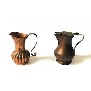 May include: Two copper jugs, one with a lid and a curved handle, the other with a straight handle and a textured body.