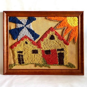 以下が含まれることがあります： 2 つの家、風車、太陽をカラフルで質感のある表現で描いた額装されたアート作品。家は黄色で赤い屋根、茶色のドアと窓。風車は青と白、太陽はオレンジと黄色です。