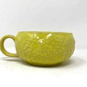 Puede incluir: Una taza de cerámica amarilla con un diseño texturizado en relieve que se asemeja a una flor o una planta. La taza tiene una forma redondeada y un asa.