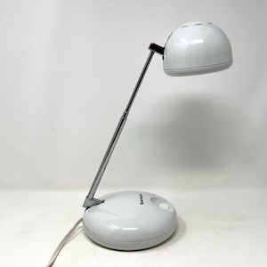 May include: A white desk lamp with a rounded base and a domed lampshade. The lamp has a chrome-colored adjustable arm and a brown accent piece. The base has a power button and a cord. The lamp is designed for desk use.
