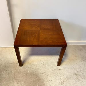 May include: A square wooden end table with a light brown finish. The table has a simple design with four legs and a flat top.