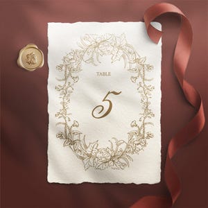 Puede incluir: Una tarjeta de número de mesa crema con un diseño de corona floral dorada y el número 5. La tarjeta tiene un borde irregular, un sello de cera dorado y una cinta roja. La palabra "TABLE" está impresa encima del número. Tema: boda.