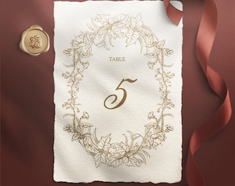 Könnte beinhalten: Eine cremefarbene Tischkarten mit einem goldenen floralen Kranz und der Zahl 5. Die Karte hat einen gezackten Rand, ein goldenes Wachssiegel und ein rotes Band. Das Wort "TABLE" ist über der Zahl aufgedruckt. Thema: Hochzeit.