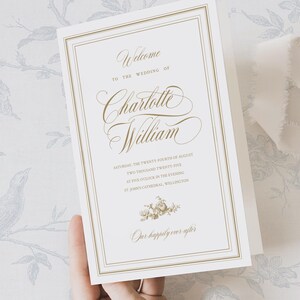 以下が含まれることがあります： 金色の文字が入ったクリーム色の結婚式の招待状。「Charlotte Williamの結婚式へようこそ」と書かれており、日付、時間、場所が記載されています。下部には小さな花の模様があり、「末永くお幸せに」という言葉が添えられています。