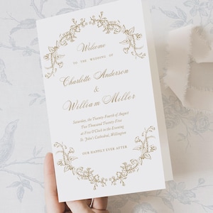 May include: A folded white wedding program with gold floral detailing and text. The program reads "Welcome to the Wedding of Charlotte Anderson & William Miller" and includes the wedding date, time, and location. The phrase "Our Happily Ever After" is also included.