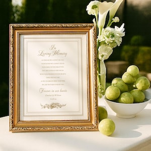 May include: A framed gold sign with the text "In Loving Memory" and a floral design. Beside it, a vase of white flowers and a bowl of green fruit sit on a white surface. The scene is outdoors with soft lighting.