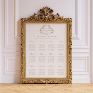 May include: A gold ornate framed seating chart with the text "PLEASE FIND YOUR SEAT". The chart lists table numbers and guest names. The frame has decorative scrollwork and is leaning against a white wall.