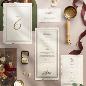 May include: An elegant wedding invitation suite with a vintage aesthetic. Includes a table number, menu, place card, and drink menu, all with gold accents and floral details. A ring box, wax seal, and ribbon add to the luxurious feel.