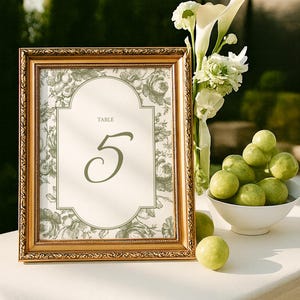 May include: A framed table number 5, with a floral design, sits on a white surface. The gold frame surrounds a cream-colored card with the word "TABLE" above the number. A vase of white flowers and a bowl of green fruit are also present.