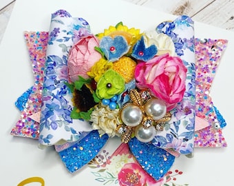 May include: A colorful floral hair bow with a blue, white, and pink floral print, a cluster of faux flowers, and a pearl and rhinestone embellishment. The bow is on a white background with the text "SimpleHearted" in gold lettering.