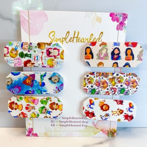 May include: Six rectangular hair clips with various cartoon character designs, including princesses and Paw Patrol characters, are displayed on a white background. The clips are white with colorful illustrations. The background includes the text "SimpleHearted" and social media information.