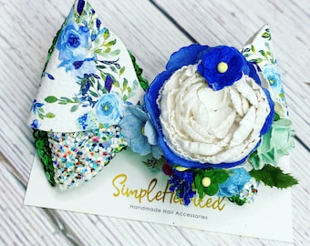 May include: A green, blue, and white floral hair bow with a white felt flower and green leaves. The bow is on a white card with the text "Simplehearted Handmade Hair Accessories".