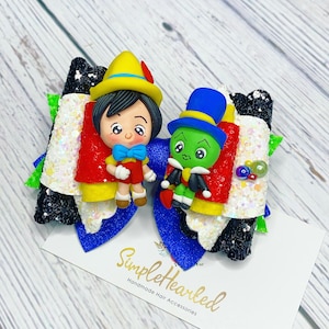 May include: A handmade hair bow featuring two character figures. One figure is a boy in red and yellow clothing, and the other is a green character in a blue hat. The bow has a blue, white, and black design with glitter accents. The text "SimpleHearted Handmade Hair Accessories" is on a card.