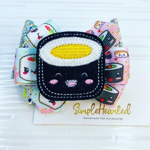 May include: A black and white sushi-shaped hair bow with a smiling face and a yellow egg on top. The bow is made of fabric and has a glittery background. The bow is attached to a white card with the text "SimpleHearted Handmade Hair Accessories".