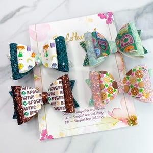 May include: Four handmade hair bows with glitter accents. The bows feature various patterns, including Girl Scout-themed designs with text and illustrations. Colors include teal, brown, pink, and white. The bows are arranged on a decorative card.