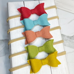 May include: Five felt hair bows in different colors, attached to nude elastic headbands. The bows are red, blue, orange, green, and yellow.