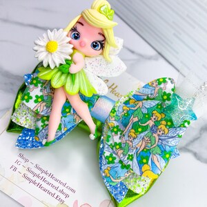 May include: A decorative hair bow featuring a fairy figurine holding a daisy. The bow has layers of green, blue, and white fabric with a glittery finish. The fairy has blonde hair and a green dress. The bow is decorated with a fairy print.